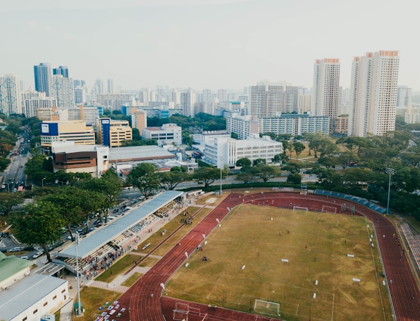 An aerial view of a sports complex with a running track surrounding a grassy field, set against the background of a cityscape. The city's skyline features a mix of tall residential buildings and shorter commercial structures. There are trees and green areas surrounding the sports facility.
