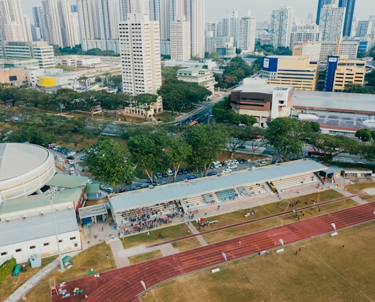 An aerial view of an urban area featuring a sports complex with a track field and a grandstand. Surrounding the sports area are numerous high-rise buildings, greenery, and roads with vehicles. The image captures people engaging in activities on the field.
