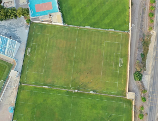 An aerial view of a sports complex featuring multiple green grass fields, including football pitches with marked goalposts. Adjacent to the fields is a blue-roofed building and a smaller area with a tennis or basketball court. Surrounding the complex, there's a road lined with trees and a dirt area with sparse vegetation.
