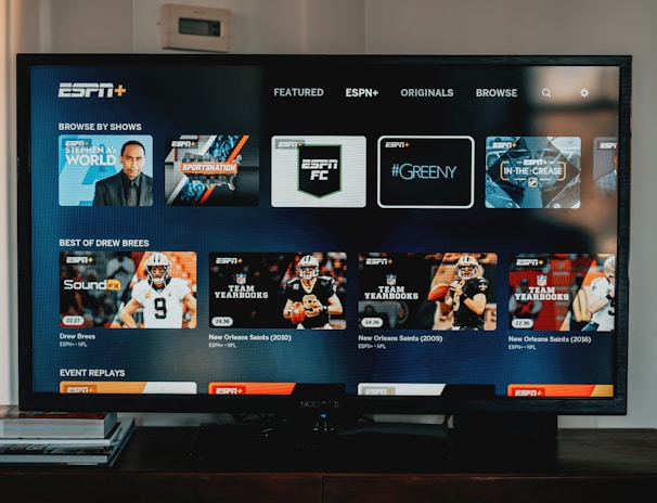 A television screen displaying the ESPN+ streaming interface with various sports-related shows and categories. The screen includes sections such as 'Browse by Shows' and 'Best of Drew Brees' with thumbnails of sports programs featuring athletes in action and analysts.