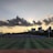 A sports field is situated in the foreground, with several people engaged in activities. The field is surrounded by a running track, and there are goalposts on either end. Behind the field, a line of modern buildings is visible, partially covering a hillside. Above, a dramatic cloud formation stretches across the sky, with the sun just below the horizon creating a silhouette effect.