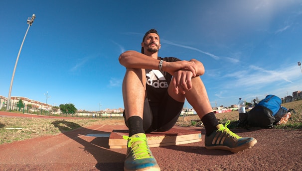 A person wearing a black athletic outfit is seated on a wooden platform in an outdoor sports field. The individual is relaxed, with arms resting on knees. The field is expansive with a clear blue sky and scattered clouds above. A brightly colored running shoe and athletic gear are visible, along with a water bottle and a blue backpack placed beside the person. Lampposts and residential buildings can be seen in the background.