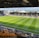 A sports stadium with a well-maintained grass field is depicted. Players are spread across the field, engaging in warm-up activities. The stands are partially filled with spectators, and the stadium is surrounded by urban buildings under a partly cloudy sky.