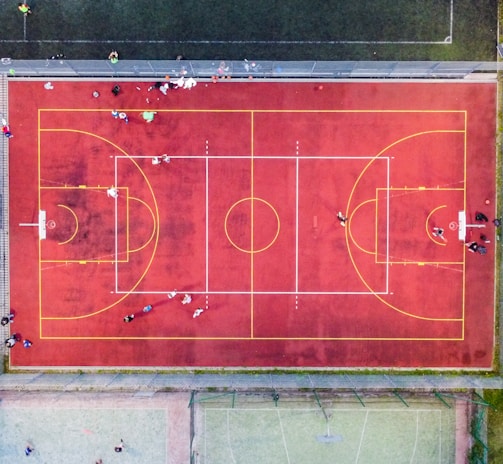 An aerial view of a sports court with a vibrant red surface and yellow lines marking different sections for basketball and soccer. Several people are present on the court, some engaged in activities, while others are standing along the perimeter. The court is enclosed by a metal fence and surrounded by grass on all sides.