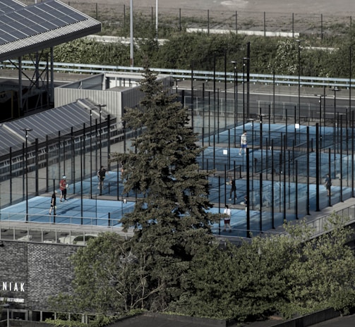 Aerial view of an outdoor sports complex featuring multiple enclosed courts with blue surfaces, surrounded by tall black fences. Several people are actively playing on the courts. Nearby is a large building with a sign reading 'MANIAK' and a tree in the foreground. Solar panels are visible on the roof of the structure beside the courts.