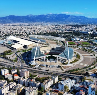 A large, modern sports complex is situated within an expansive urban landscape. The main stadium features prominent architectural structures and an open roof design. Surrounding the stadium are various sports facilities, roads, and greenery. Residential and commercial buildings extend into the distance, framed by mountain ranges under a clear blue sky.