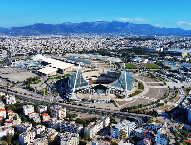 A large, modern sports complex is situated within an expansive urban landscape. The main stadium features prominent architectural structures and an open roof design. Surrounding the stadium are various sports facilities, roads, and greenery. Residential and commercial buildings extend into the distance, framed by mountain ranges under a clear blue sky.
