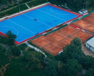 An aerial view of a large sports complex with a vibrant blue field surrounded by a red boundary and adjacent clay courts. The complex is nestled amidst dense greenery and trees, with a few buildings visible in the background. The setting appears to be serene and well-maintained, with a sense of spaciousness.