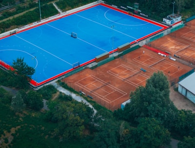 An aerial view of a large sports complex with a vibrant blue field surrounded by a red boundary and adjacent clay courts. The complex is nestled amidst dense greenery and trees, with a few buildings visible in the background. The setting appears to be serene and well-maintained, with a sense of spaciousness.