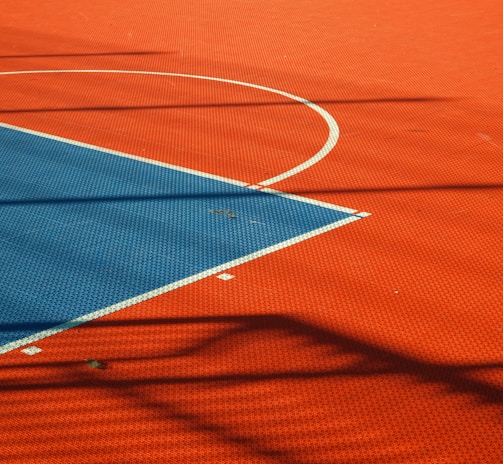 A vibrant sports court with sections of red and blue, featuring crisp white lines marking the boundaries. Shadows from a structure create geometric patterns across the surface, adding depth and interest to the scene.