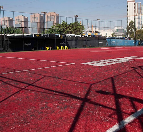 A sports court with a red playing surface and white lines, surrounded by a black mesh fence. In the background, tall buildings are visible, along with some trees and a few yellow chairs positioned near the fence.